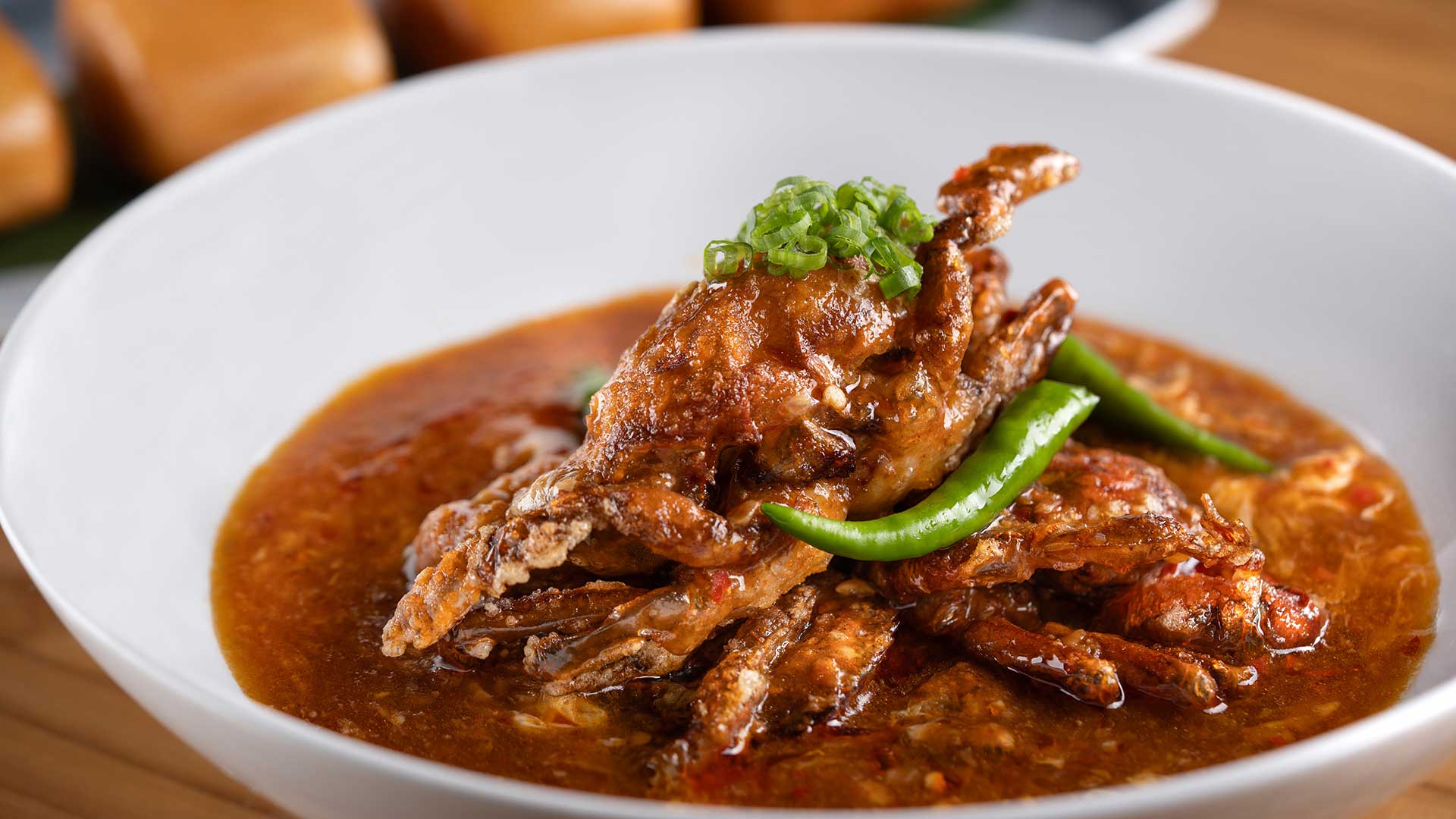 A white bowl of crab curry garnished with green chilies and chopped green herbs, with bread rolls in the background.