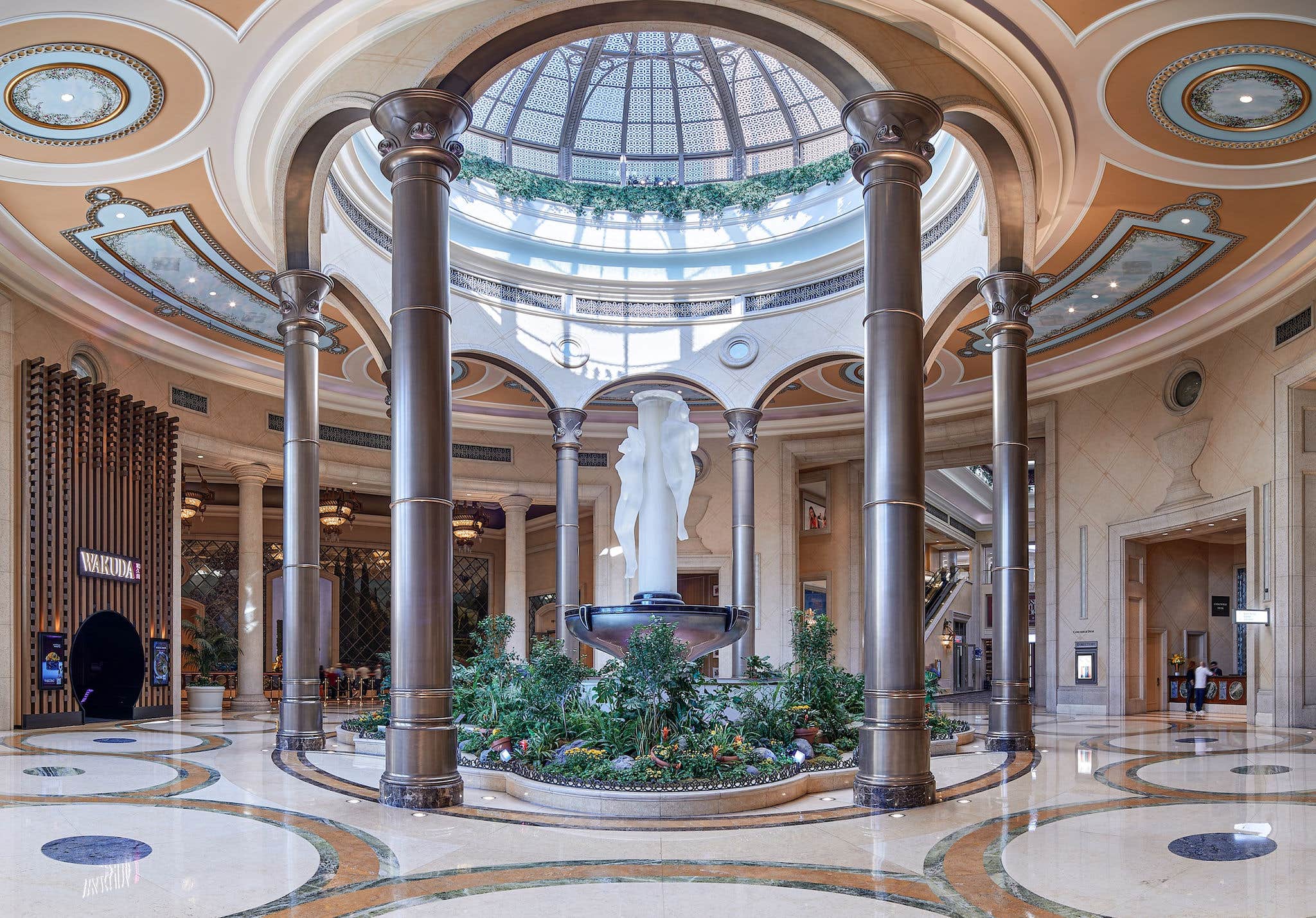 Luxurious lobby with marble floors, a central statue, tall columns, and a large domed glass ceiling.