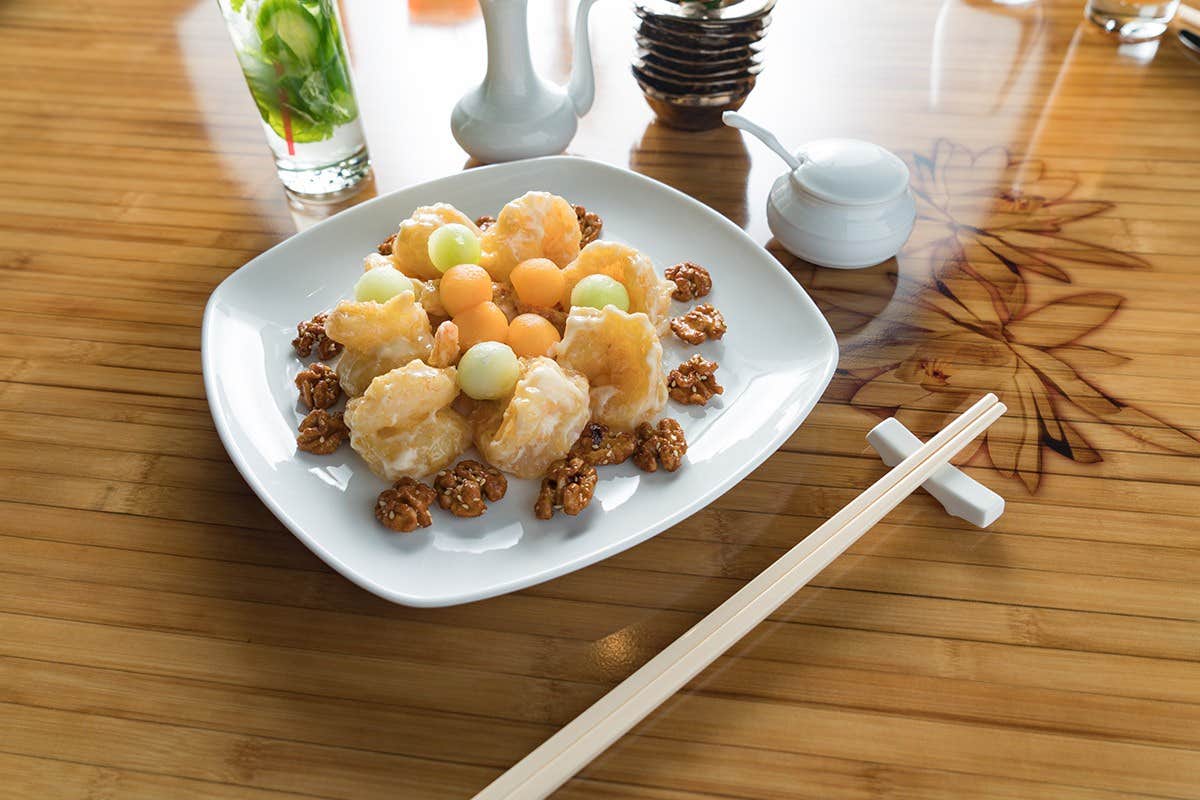 A dish with fried shrimp and melon balls on a white plate, garnished with walnuts, next to chopsticks and a drink.