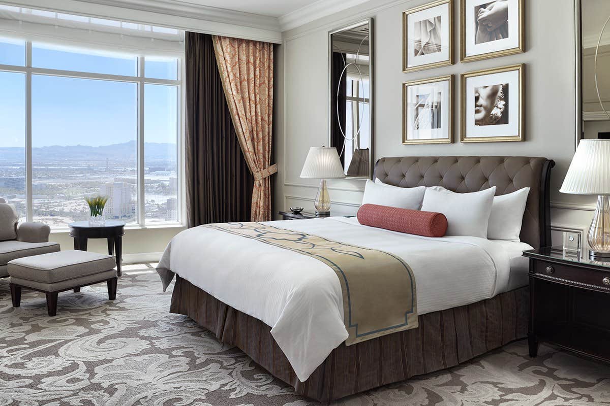 Elegant hotel room with a large bed, stylish decor, a window view of a cityscape, and armchairs near the window.