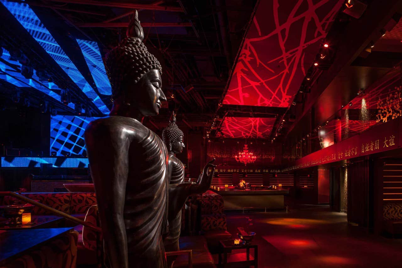 Dimly lit club interior with Buddha statues, red and blue lighting, and abstract ceiling patterns.