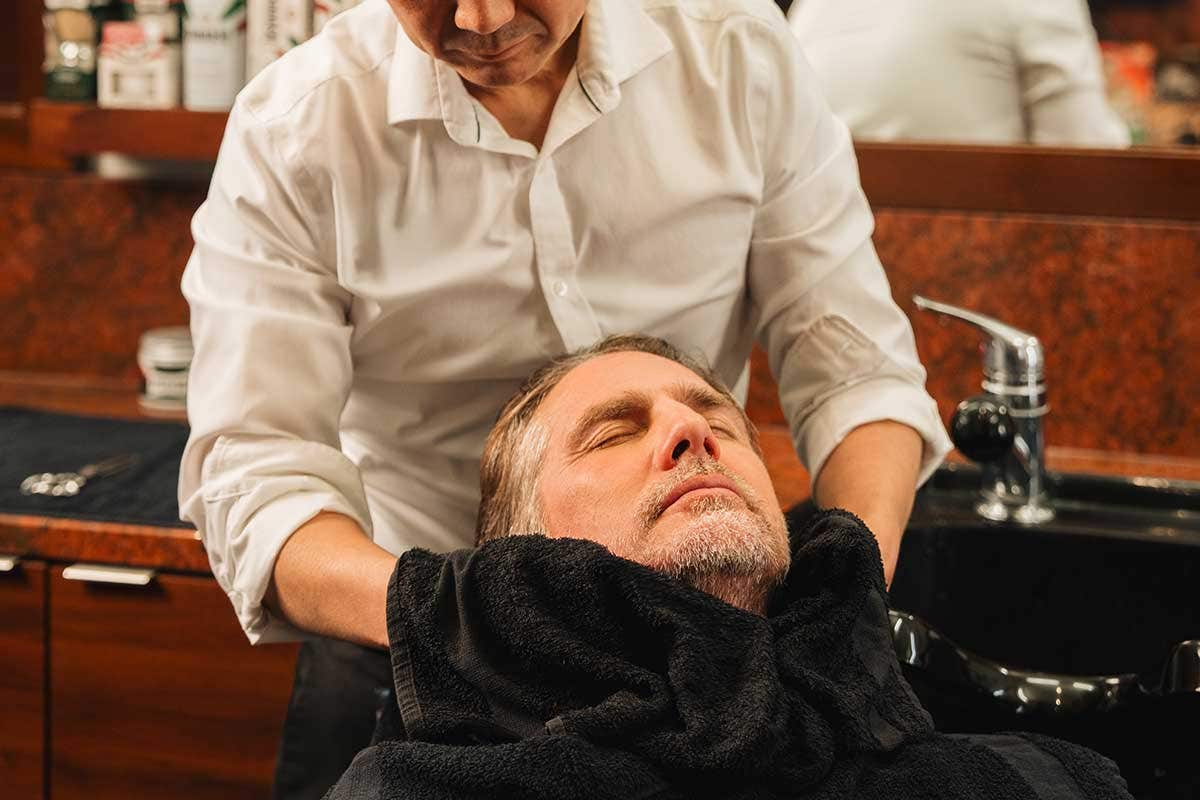 A man reclines in a salon chair with head tilted back while another person in a white shirt wipes the man’s beard with a dark towel as part of a barber beard trim and shave service; bottles and a mirror are visible on a wooden counter in the background.