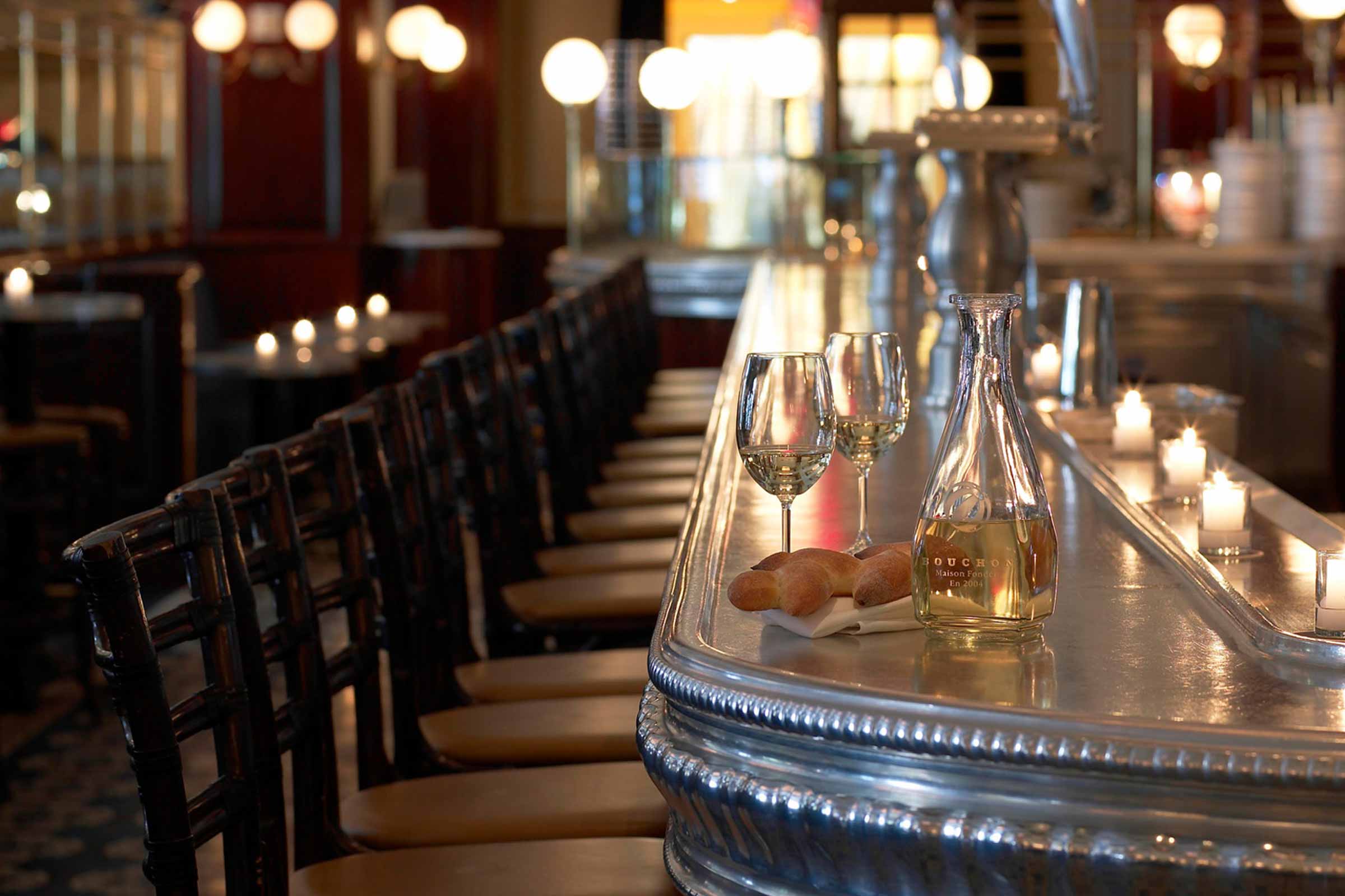 Elegant bar with empty seats, wine glasses, a wine bottle, and breadsticks on a polished countertop, lit by ambient lamps.