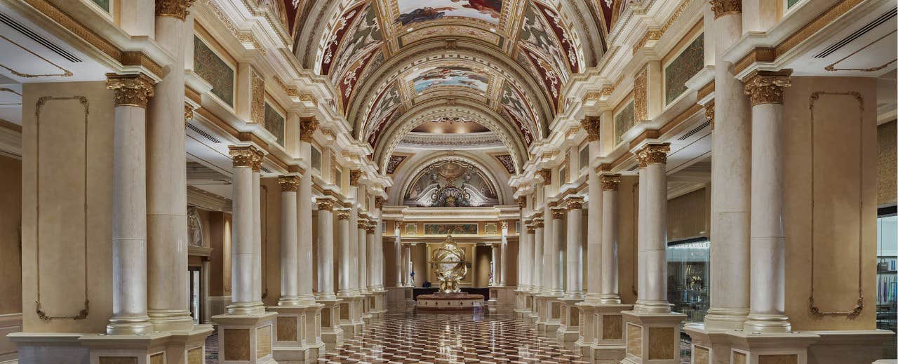 A grand, ornate hallway with tall marble columns, detailed ceiling murals, and a central gold statue at the end.