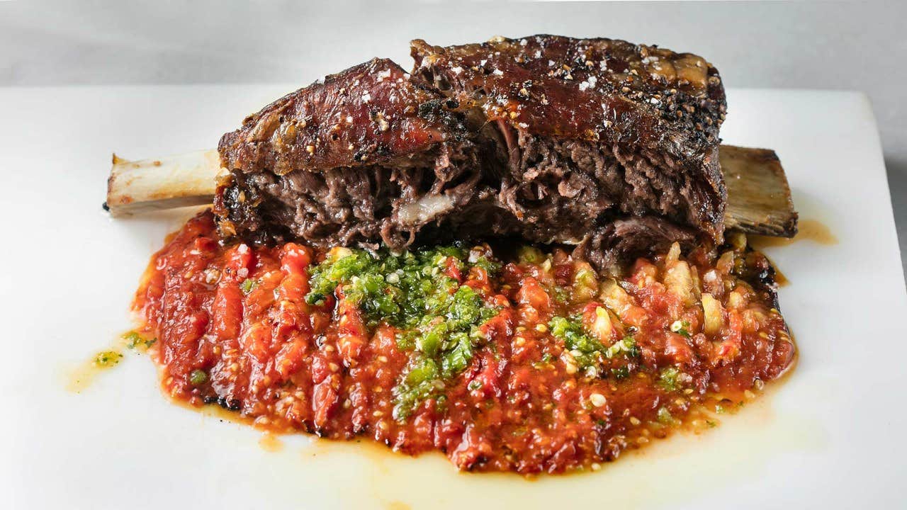 A cooked beef short rib with a side of red and green salsa, served on a white plate.