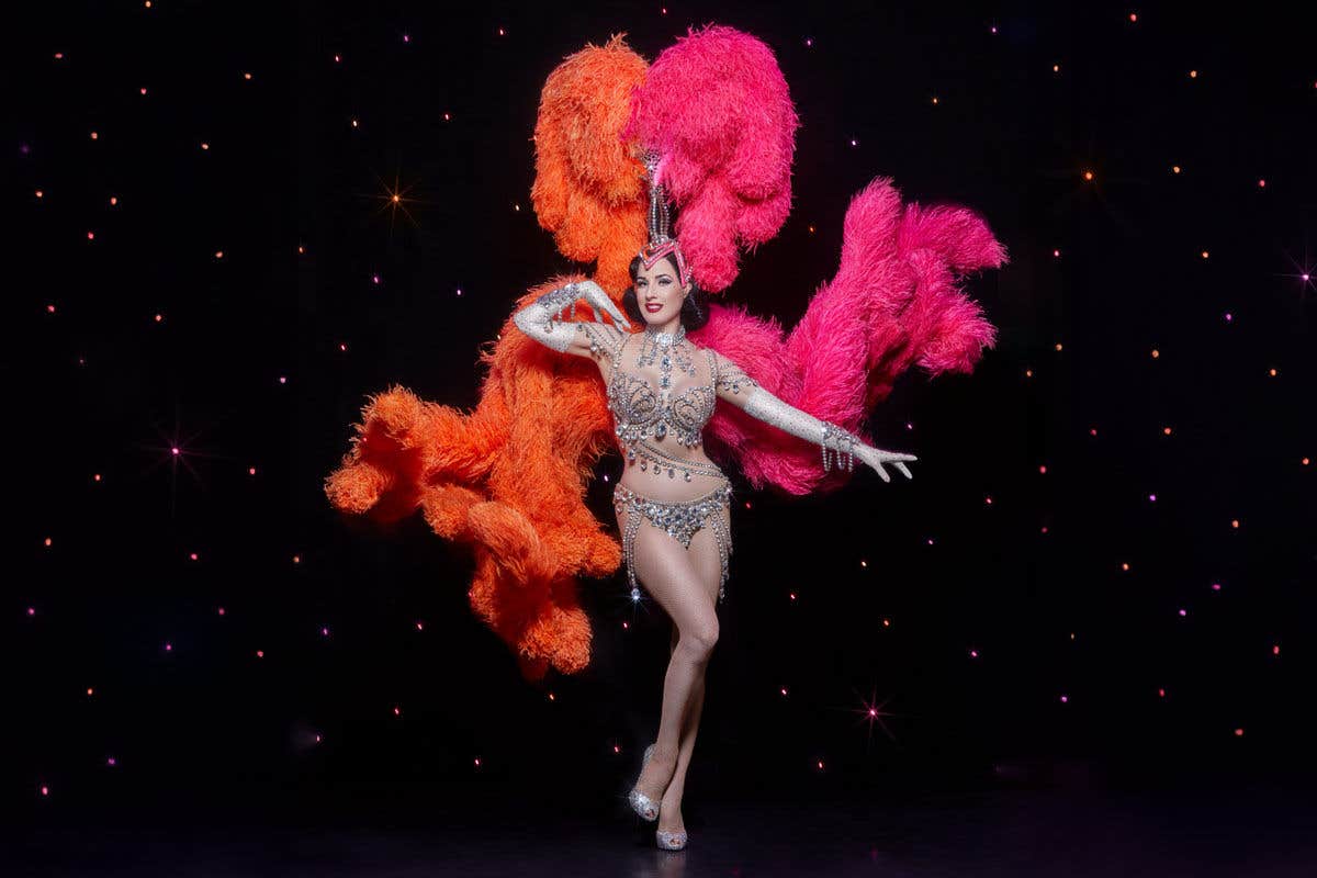 Dita Von Teese in a diamond-covered outfit with an orange and pink headdress in front of a starry backdrop