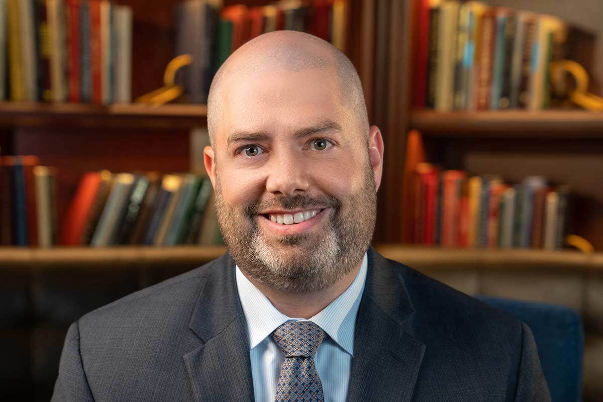 A man in a suit with a beard and shaved head smiles in front of a blurred bookshelf background.