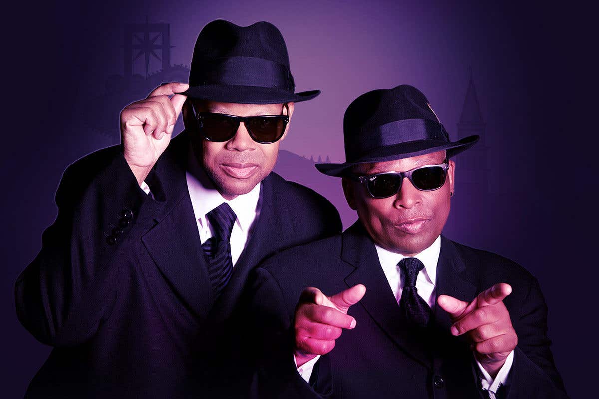Jimmy Jam and Terry Lewis in black hat, dark sunglasses, white shirt, black tie and black jacket looking at the camera.