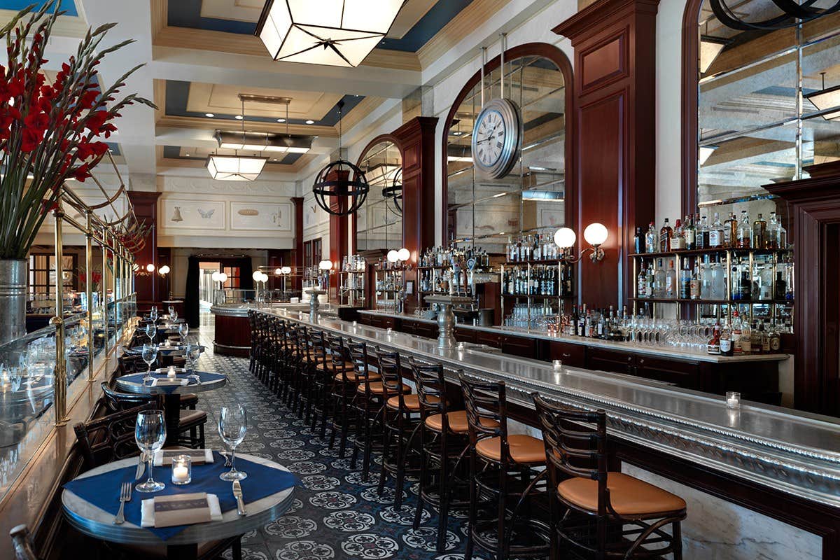 Elegant restaurant bar with high stools, a long counter, decorative tiles, and a clock on the mirror behind the bar.