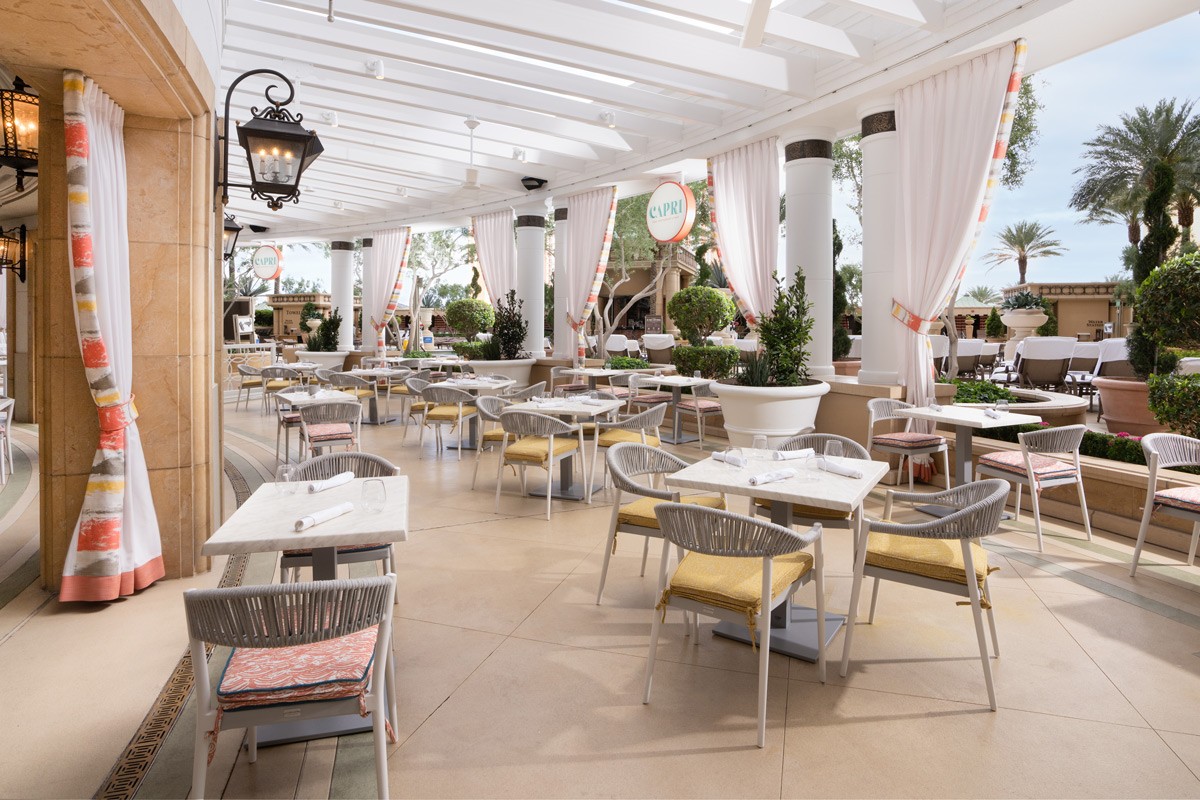 An elegant outdoor restaurant with neatly arranged tables and chairs under a white pergola, framed by lush green plants.