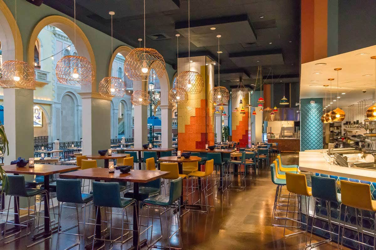 A modern restaurant with colorful seating, arched windows, and hanging woven lamps over tables and a bar area.