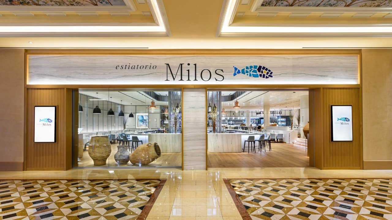 Entrance of Estiatorio Milos restaurant with modern decor, showcasing logo and interior seating through large glass doors.