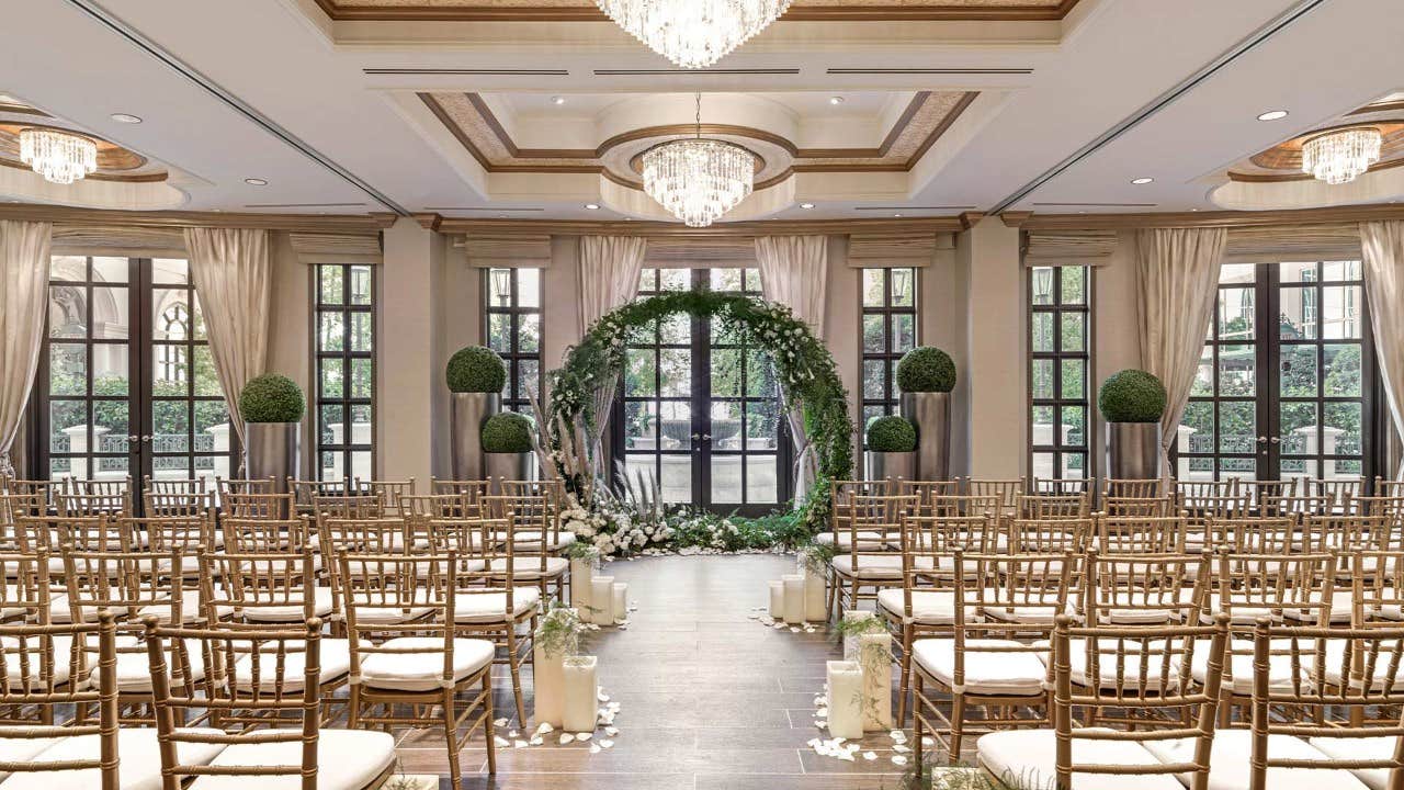 Elegant wedding venue with chandeliers, greenery, white flowers, and rows of chairs facing a floral arch in front of windows.