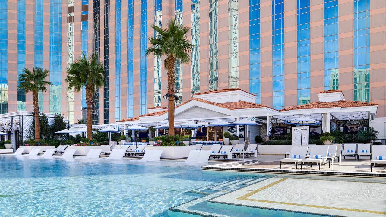 A pool with lounge chairs and umbrellas, surrounded by tall buildings and palm trees in a resort setting.