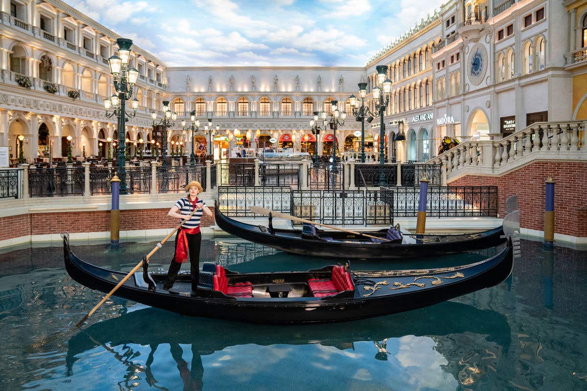A gondola navigates an indoor canal surrounded by Italian-themed buildings with bright, clear skies above.