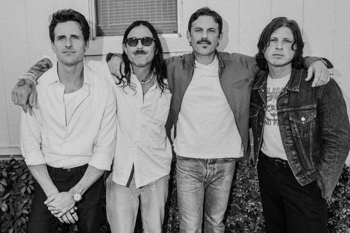 A grayscale photo of Kings of Leon band members standing together in front of a building looking directly at the camera. 