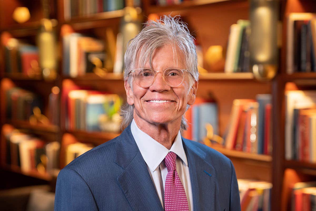 An elderly individual with glasses and a suit smiling in front of a bookshelf background.