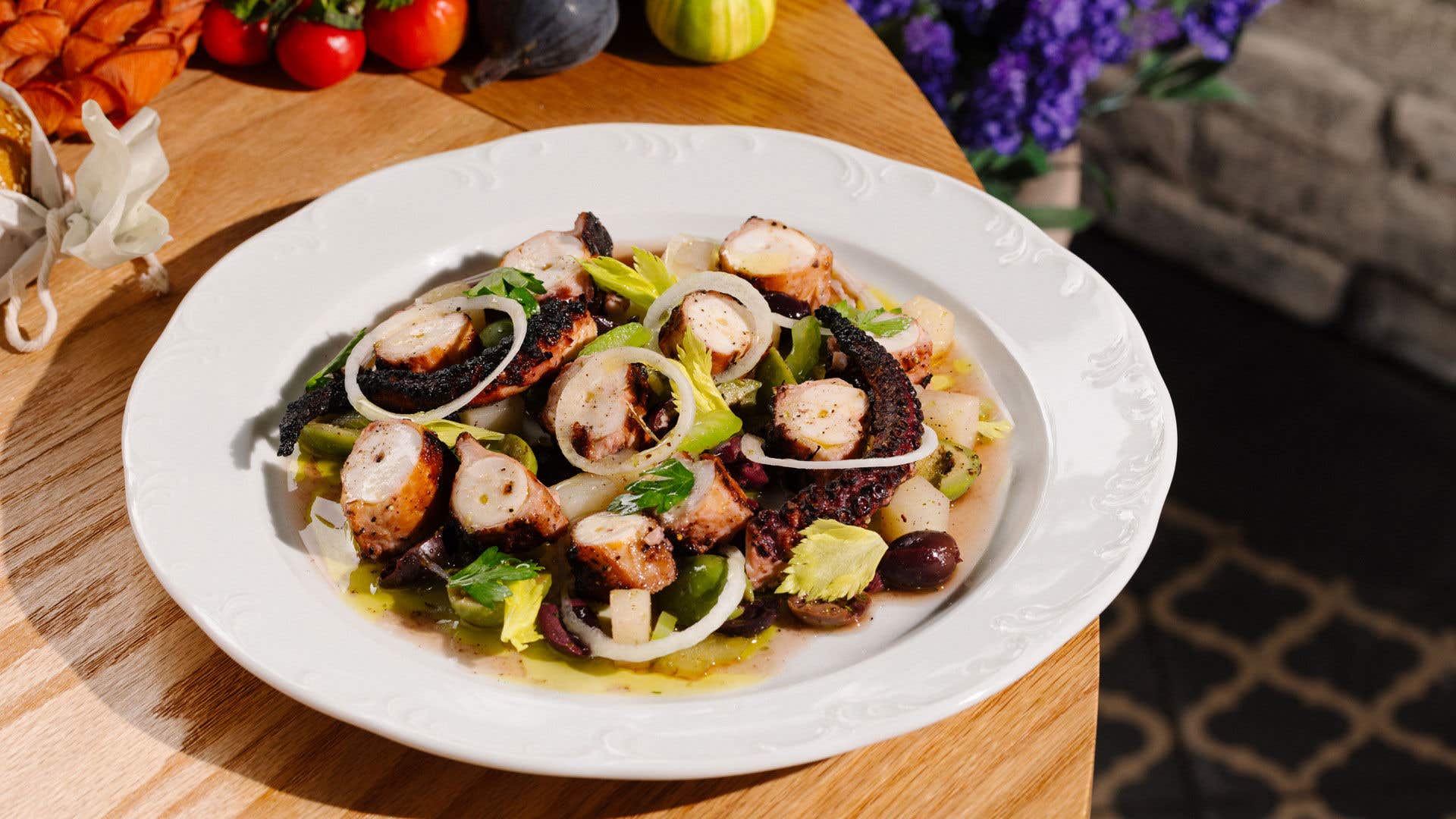 Octopus dish on a white plate on a wood table with assortment of vegetables nearby.