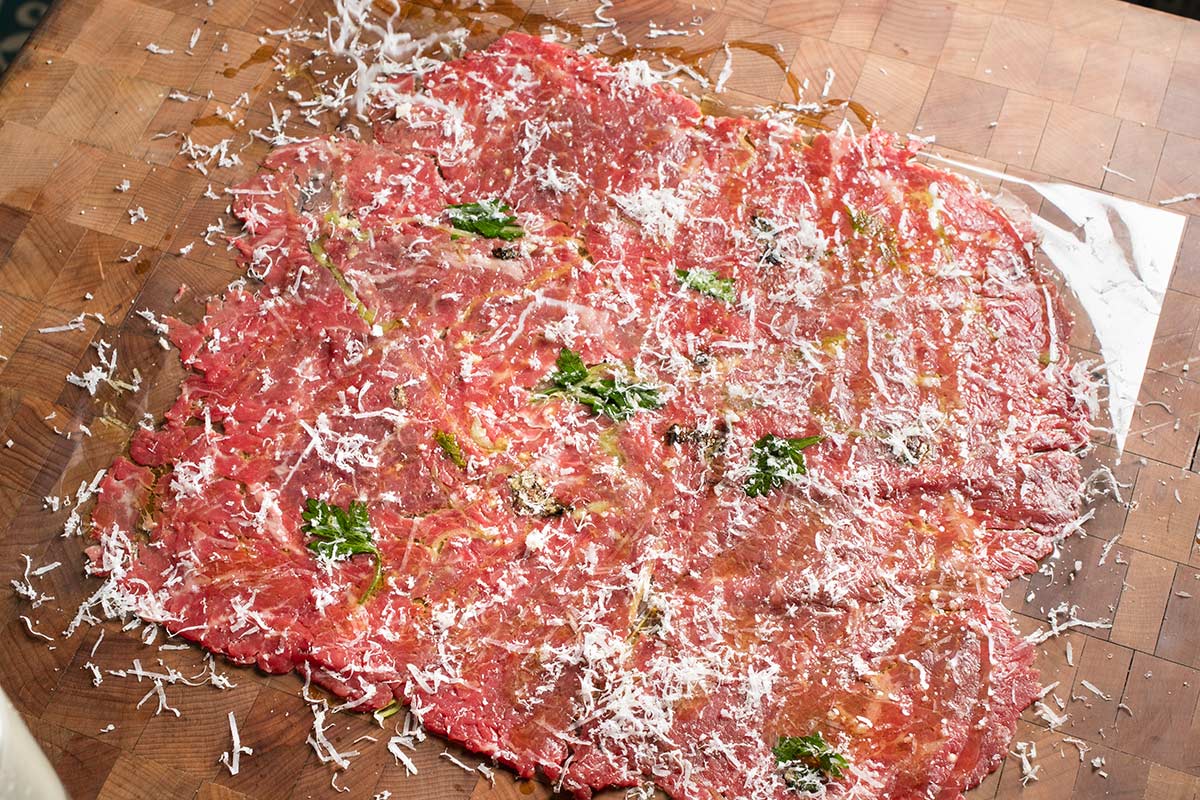 Thinly sliced raw beef carpaccio topped with grated cheese, herbs, and olive oil, on a wooden cutting board.