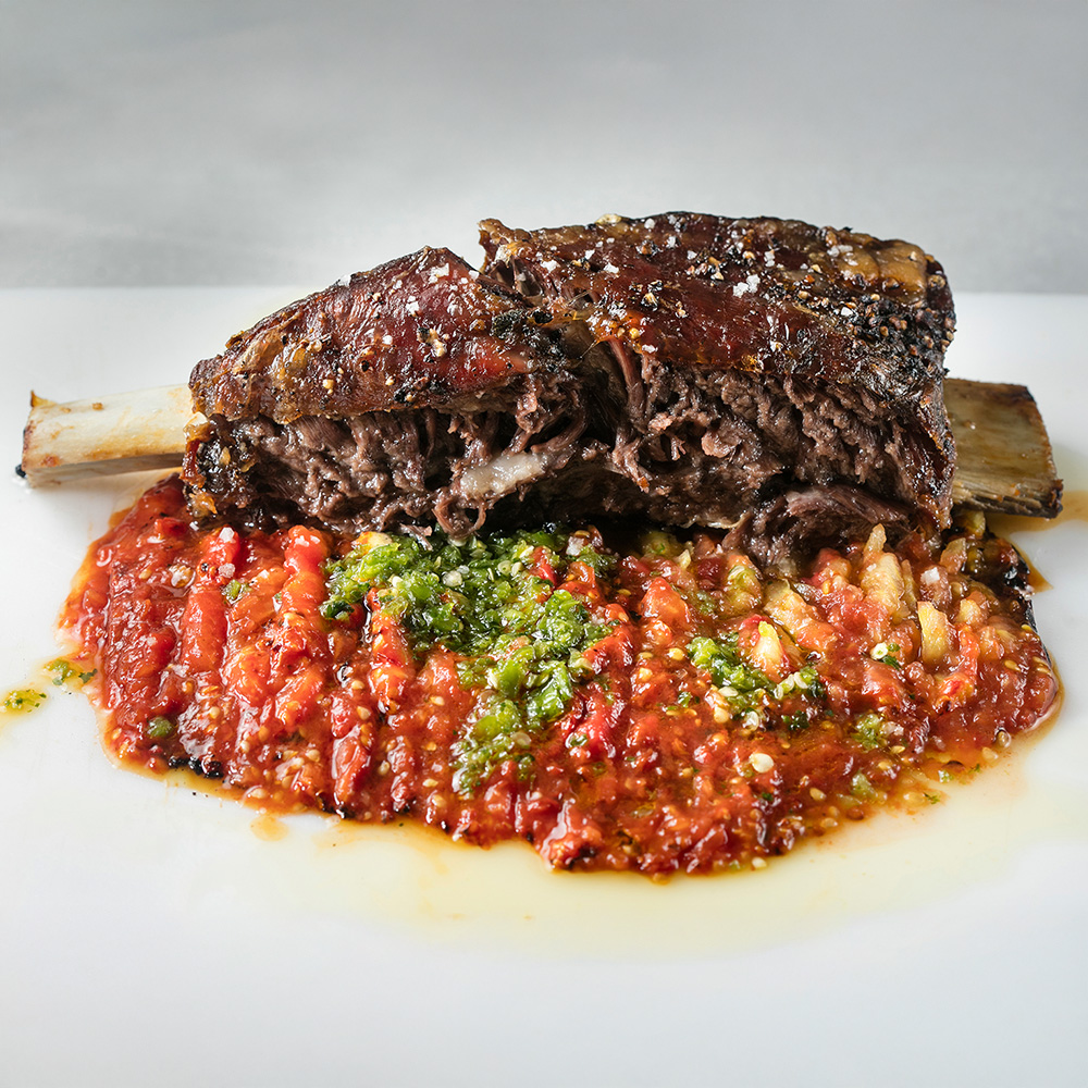 A cooked beef short rib with a side of red and green salsa, served on a white plate.