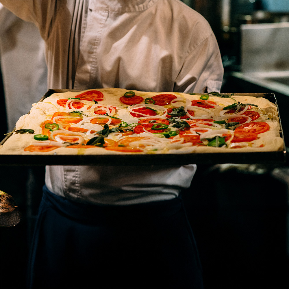 A chef holds a rectangular tray of uncooked pizza topped with tomatoes, onions, and green peppers.