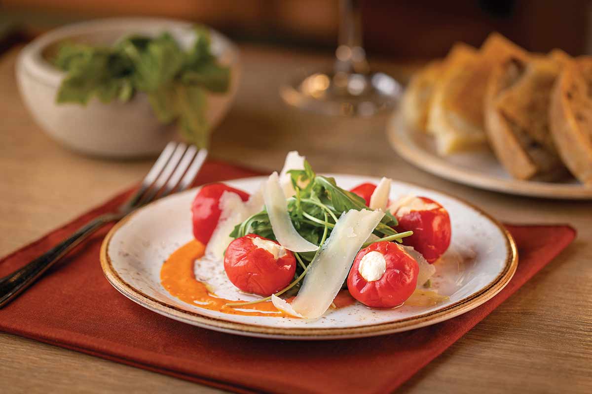 A plate of stuffed cherry peppers with arugula, shaved cheese, and a red sauce, served on a table with bread and greens.